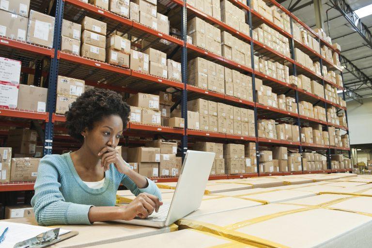 African American female warehouse worker working on inentory on a lap top computer in a large distribution warehouse of products stored in cardboard boxes and on large racks.